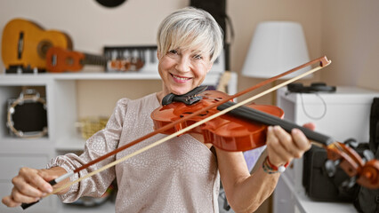 Smiling mature woman with grey hair playing violin indoors with musical instruments background. © Krakenimages.com