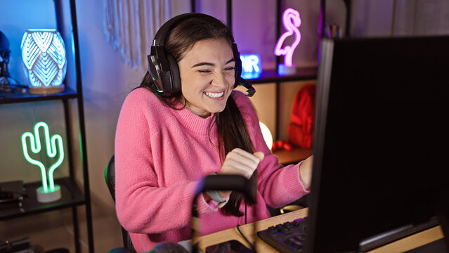Hispanic woman gaming indoors with headphones, steering wheel, and neon lights at night. - Powered by Adobe