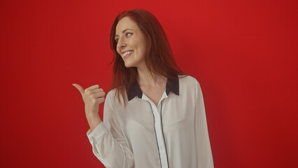 A smiling young adult caucasian woman with red hair, striking a pointing pose over an isolated red background.