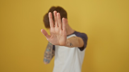 A young man with a tattoo gesturing stop against a vibrant yellow backdrop, portraying a message of boundaries.