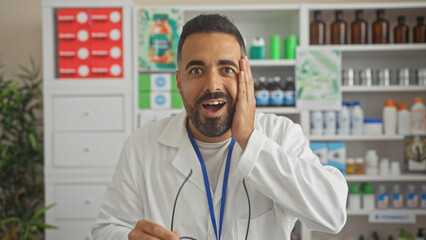 Surprised hispanic man in white lab coat holding glasses inside a pharmacy with shelves full of products.