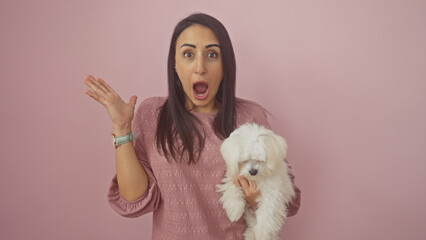 A shocked hispanic woman holds a white bichon maltese against a pink wall, conveying surprise and companionship.