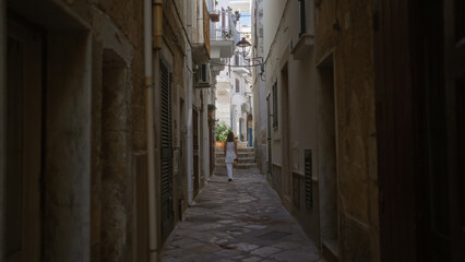 Fototapeta premium A young, beautiful, hispanic woman walks down a charming narrow street in polignano a mare, puglia, italy, showcasing the quaint architecture and mediterranean atmosphere.