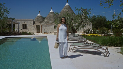 A beautiful young hispanic woman stands by a sparkling pool next to traditional trulli houses in...