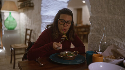 Young hispanic woman enjoying a meal indoors at a cozy italian restaurant, focused on eating with a fork, surrounded by warm, rustic decor and casual ambiance.