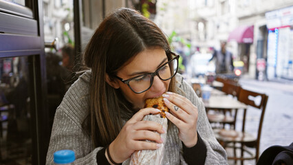 Adult hispanic woman enjoys a kebab on a european city street, suggesting tourism and urban dining.