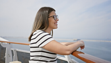 Smiling young woman leaning on a cruise ship railing overlooking the ocean under a clear blue sky.