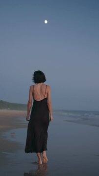 A young woman with short hair, wearing a long flowing dark dress, walks barefoot on the beach as the moonlight shines on the water behind her.