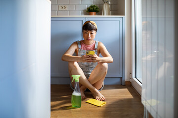 Young woman cleaning kitchen while checking her smartphone at home