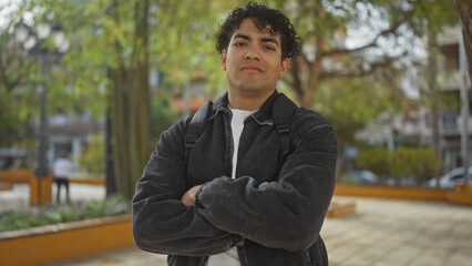 Obraz premium Young hispanic man with crossed arms standing confidently in an urban park setting.