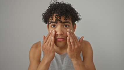 Handsome young hispanic man with curly hair touching his face, isolated against a white wall