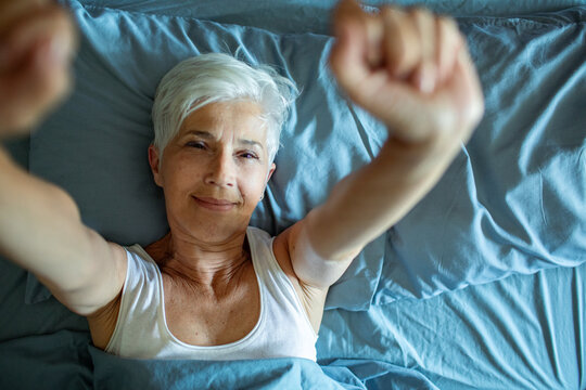 Senior woman waking up in bed and stretching arms with a smile