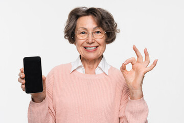 Smiling positive elderly woman grandmother in glasses hold in hand use mobile cell phone with blank screen workspace area show ok gesture isolated on white studio background