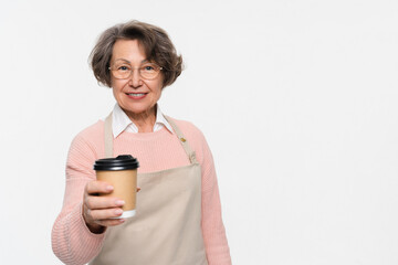 Fototapeta premium Smiling cheerful mature elderly woman female barista wear beige apron work in shop hold give craft paper brown cup coffee to go isolated on light white background. Small business startup concept