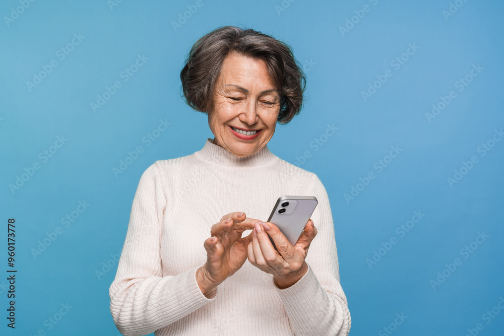 Wall mural Happy aged caucasian woman grandmother holding smartphone using mobile online apps, browsing. Smiling old elderly lady texting sms message chatting with family on phone isolated over blue background.