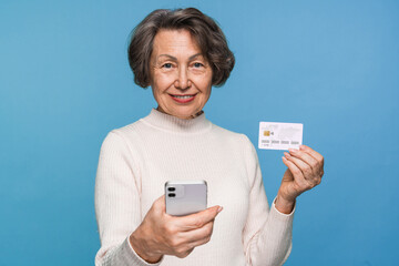 Smiling beautiful elderly old mature woman grandmother in casual clothes holding using mobile cell smart phone and credit bank card, standing isolated on blue background. Online shopping concept