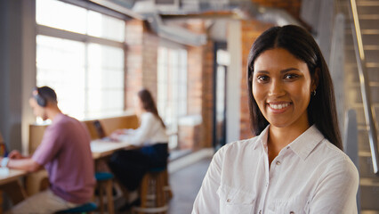 Portrait Of Young Smiling Businesswoman Working In Open Plan Office With Colleagues In Background