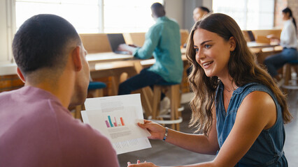 Two Young Businesspeople Meeting In Office Discussing Documents With Colleagues In Background