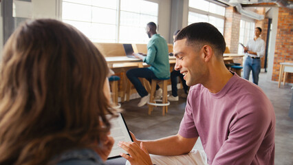 Two Young Businesspeople With Digital Tablet Meeting In Office With Colleagues In Background