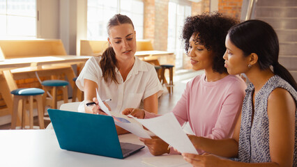 Three Young Businesswomen Meeting In Modern Office Sitting Around Table Working On Laptop Together