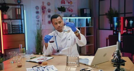 Man doctor chemist in uniform working on laptop and viewing chemical solutions in flasks during his work in evening lab