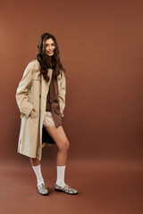 A young woman exudes confidence in a stylish autumnal outfit, posing effortlessly against a warm brown backdrop.