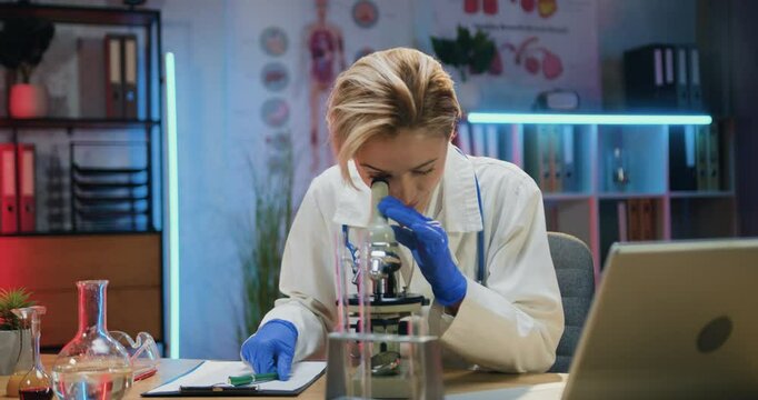 Woman doctor chemist in white coat and sterile gloves noting important details into report while viewing samples under microscope
