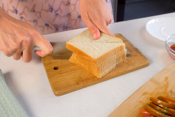Woman cutting toast bread on wooden cutting board cooking hot sandwiches on kitchen at home. Preparing snack fast food for herself. Ingredients tomatoes, sausage, sauce on table.