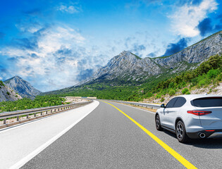 Fototapeta premium A white car drives along the highway against the backdrop of rocky mountains on a sunny day.