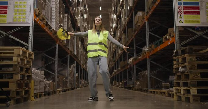 Warehouse worker dancing joyfully in safety vest and casual jeans amidst tall shelves and packed pallets in a spacious, high-ceilinged distribution center