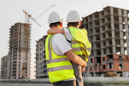 Father and son in protective helmets and vests look at the construction of a building at sunset.