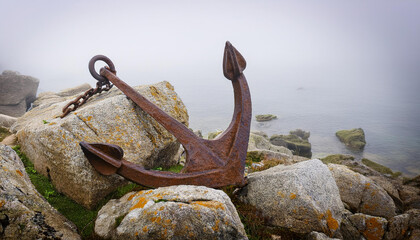 Rusty anchor among weathered rocks at foggy seaside, maritime history.