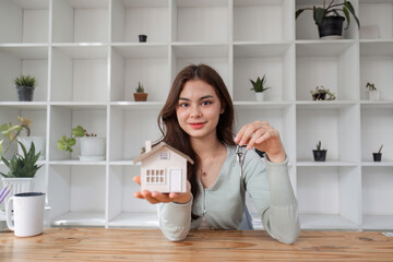 Smiling Real Estate Agent Holding House Model and Keys in Modern Office with Shelves and Plants