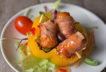 Close-up of Bacon Wrapped Sausage With fresh vegetables of various colors on white plate.