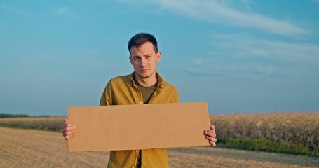 A man holds a blank cardboard sign while standing in a field, wearing a yellow shirt with a clear...