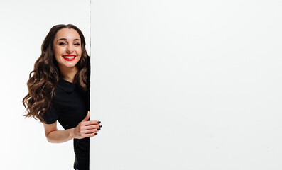 Cheerful woman with long hair peeks from behind a white wall. A smiling woman with long, wavy hair...
