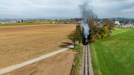 A steam locomotive chugs along railway tracks adjacent to expansive farmland and trees during autumn.