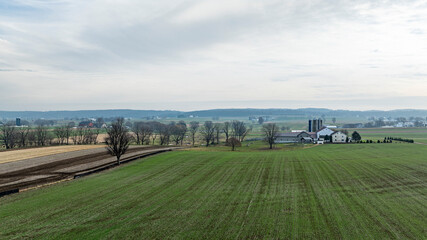 Fototapeta premium Rolling green fields stretch out in early spring, while a quaint farmhouse sits among trees. Distant hills provide a peaceful backdrop, creating a serene rural atmosphere.