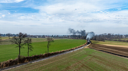 A steam locomotive chugs along a winding track surrounded by lush green fields and distant trees. The sky is partly cloudy, adding a serene atmosphere to the springtime scenery.