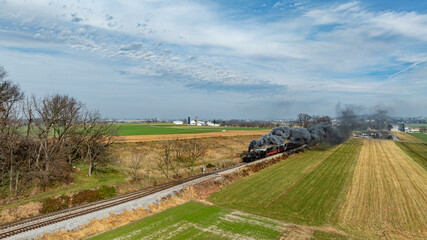 A vintage steam locomotive chugs along the railway surrounded by expansive green fields and farms on a bright and clear day, emitting plumes of grey smoke into the sky.