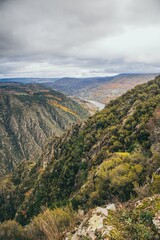 The image shows a rugged, mountainous landscape with a deep valley and a meandering river. The rocky terrain and sparse vegetation lead to steep cliffs. Overcast skies suggest cool or cloudy weather.