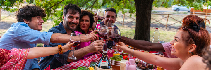 happy friends toasting red wine glasses at barbecue table in countryside – group of young people having fun bbq dinner party – making barbeque in backyard dinner time - horizontal web banner size for 