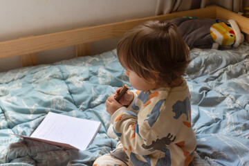 Bebé de un año sentado en su cama, él está pintando con sus colores sobre un cuaderno con hojas...
