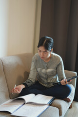 Focused young asian teen girl student pupil writing notes in notebook distance learning studying online makes goals check list on sofa at home. Remote college school education, e learning concept.