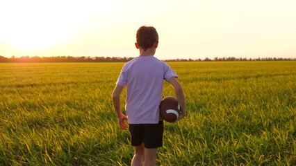 boy running on a green meadow against the background of a beautiful sunset. a child boy run on the grass in the park holding a rugby ball. child dream