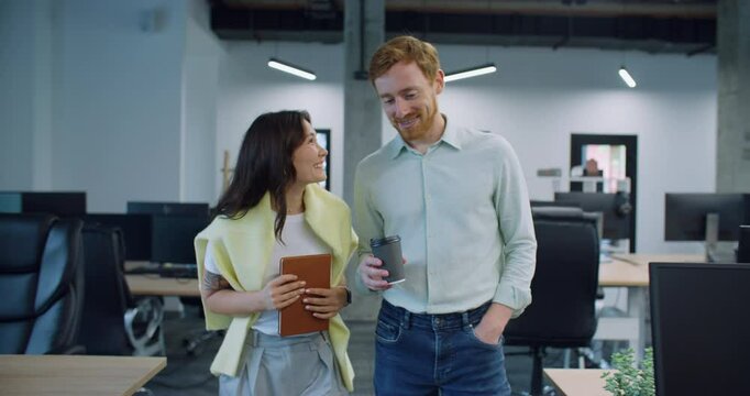 Cute couple walking through their workspace. Casually talking and smiling while enjoying conversation. Discussing possible plans for evening during coffee break. Modern room. Many desks with monitors.