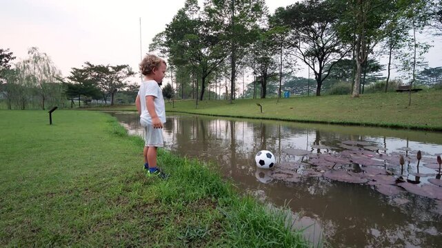 Toddler stands by edge of creek in park, staring at soccer ball floating just out of reach in water. Little boy is puzzled and frustrated as he watches ball drift, unsure how to retrieve it.