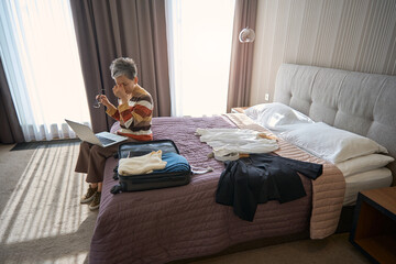 Tired senior woman working with laptop sitting on the bed in hotel room