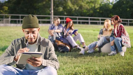 An African American student in a khaki shirt and a stylish hat works on his homework in the foreground, while his cheerful classmates are seated in the background at a university park