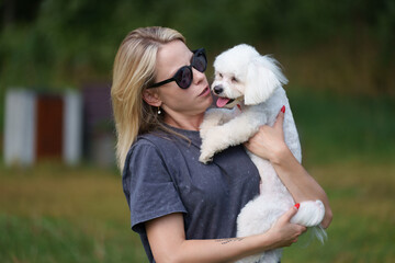 Portrait of smiling young woman kiss dog in Natural Park. Dog lover woman wearing blue dress with dog during good day. Concept of friendship and beautiful girl with a small dog